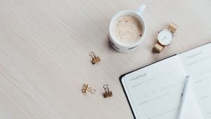 A minimalist desk arrangement featuring a coffee cup, notebook, pen, and wristwatch.