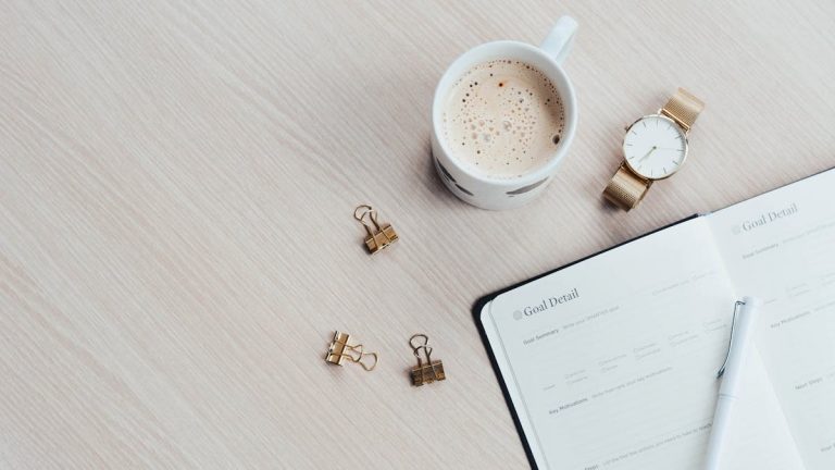 A minimalist desk arrangement featuring a coffee cup, notebook, pen, and wristwatch.